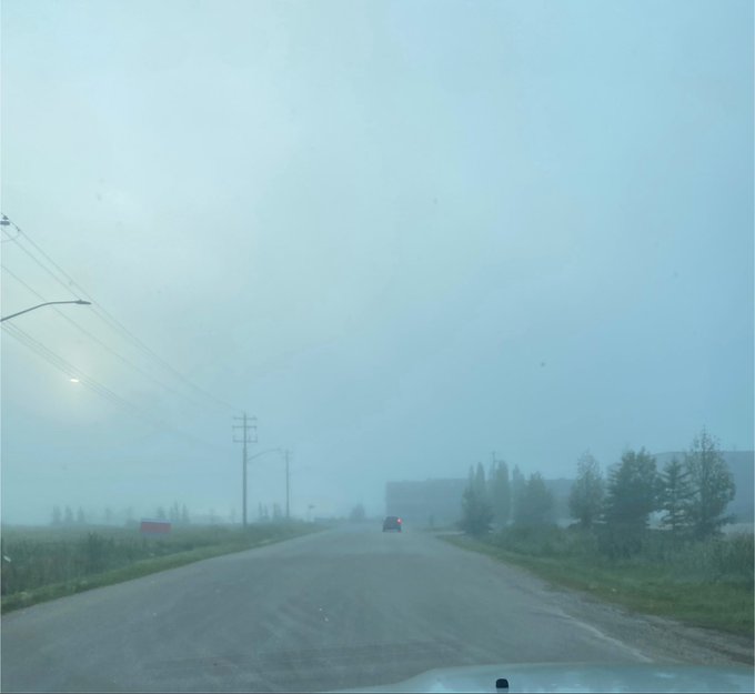 A view from inside a vehicle driving down a foggy, misty road with power lines on the left and a car with red taillights ahead.