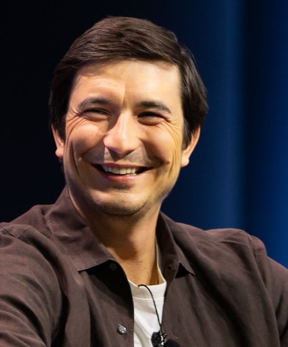 A man in a dark button-up shirt smiles broadly at what appears to be a conference or panel event. The image accompanies a post about Robinhood expanding trading hours and launching new perpetual futures products, suggesting he may be a Robinhood executive discussing these developments.