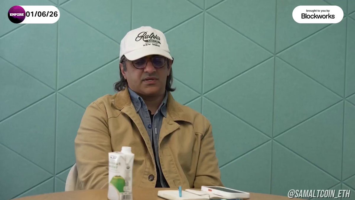 The image features a man wearing sunglasses, sitting at a table with a book open in front of him. The background shows a green wall, which contrasts with the man's tan jacket. There are two other people visible in the image, one on the left side and another person partially out of frame on the right side. A bottle is also present near the center of the scene.