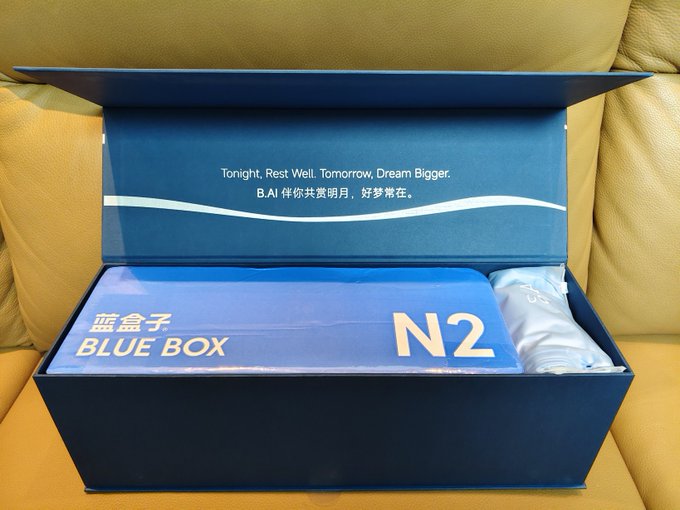 An open blue 'BLUE BOX N2' package on a tan leather couch, with the inside lid reading 'Tonight, Rest Well. Tomorrow, Dream Bigger. B.AI 伴你共赏明月, 好梦常在.'