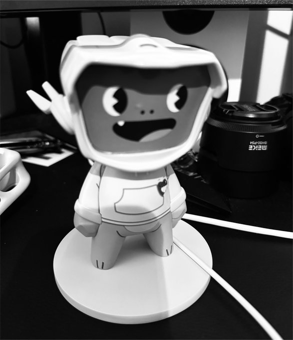 A black-and-white photo of a Blokyz collectible figure with a large open-mouthed smile, wearing a hoodie, displayed on a white circular base. Visible text on a camera lens in the background reads "SIGMA 30mm" and "DC DN," adding a sense of an at-home collector's setting, which aligns with the discussion about collectibles.