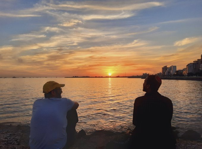 Silhouette of two people sitting by the water at sunset, with a city skyline visible in the distance across the bay.