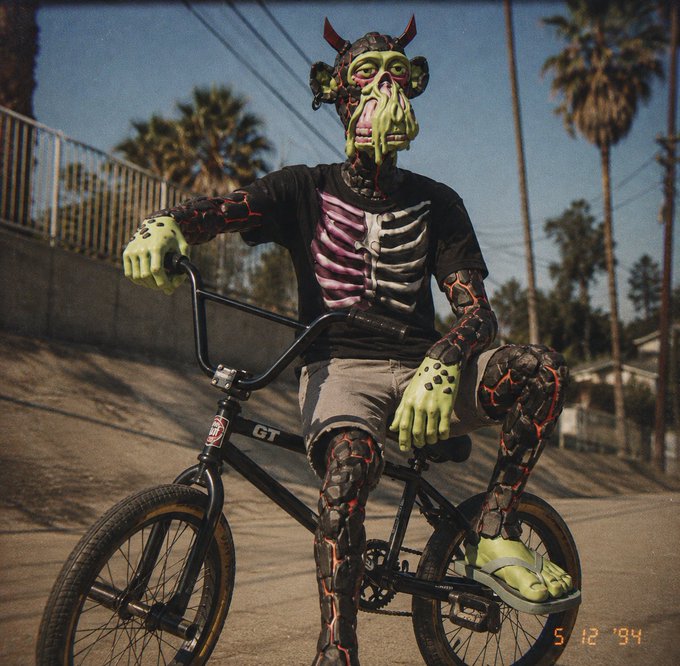 A person in elaborate green-skinned devil and skeleton-themed prosthetic makeup rides a GT BMX bike down a palm-tree-lined street, with a '5 12 '94' timestamp visible.