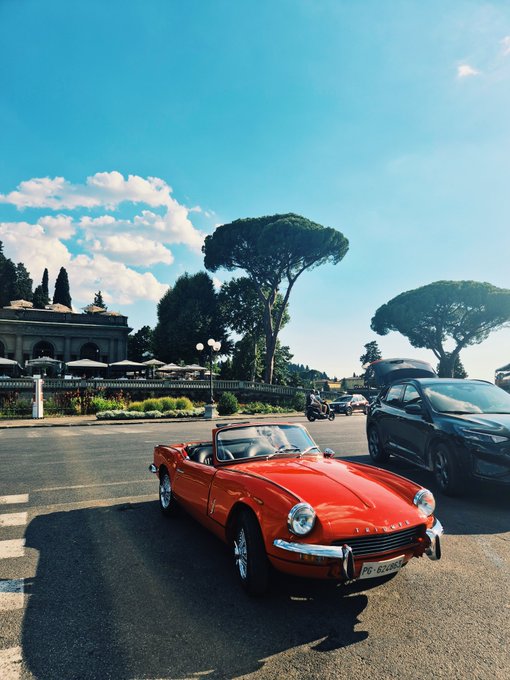 A vibrant red classic convertible, likely a Triumph Spitfire, is parked on a sunlit street with Italian umbrellas and Mediterranean pine trees in the background, displaying a license plate reading "PG 522592." The scene evokes a leisurely Italian morning, fittingly complementing the cheerful "Good Morning #ShibArmy" greeting.