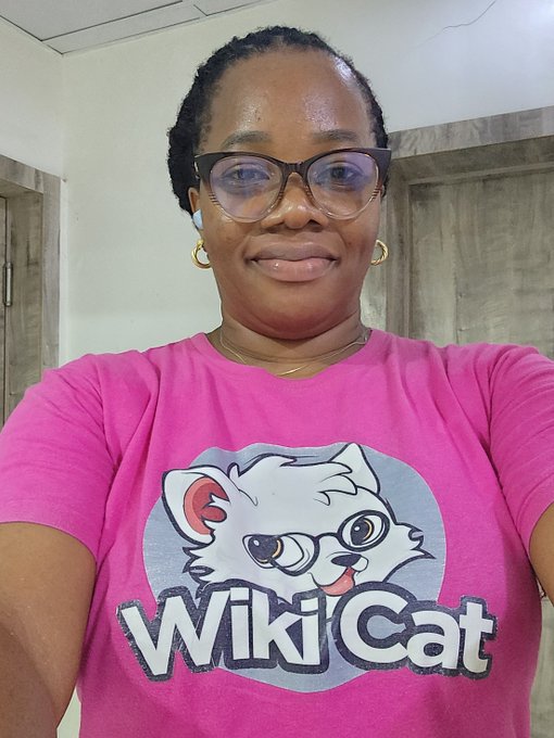 A smiling woman wearing glasses and a pink "WikiCat" t-shirt poses for a selfie, representing her support for the $WKC cryptocurrency token as part of a bullish investment message.
