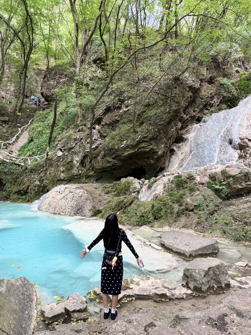 A woman with long dark hair wearing a black polka dot dress stands on rocks looking out at a turquoise blue pool and waterfall surrounded by lush green forest.