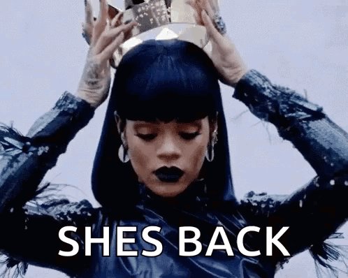 The image features a woman wearing a crown of gold and black feathers, with her arms raised high above her head. She has dark hair styled in an updo and is wearing a blue outfit that includes a skirt and top. The caption below the image reads "She's back", which could be interpreted as a reference to the song "She's Back" by the artist Radiohead.