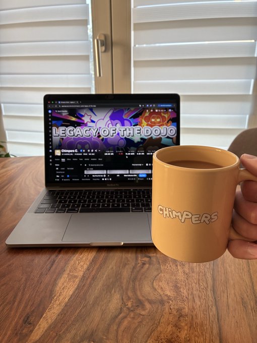 A hand holding a tan 'CHIMPERS' branded mug filled with coffee, with a laptop displaying the 'LEGACY OF THE DOJO' YouTube channel by Chimpers in the background.