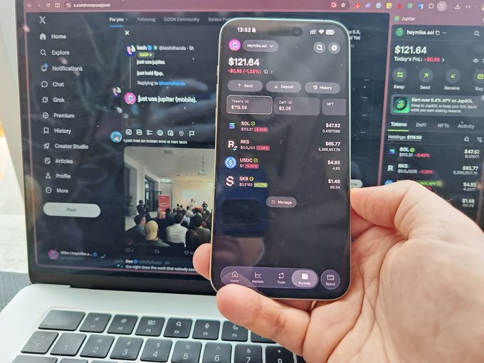 A hand holding a smartphone displaying the Jupiter crypto portfolio app showing a balance of $121.64, with a laptop in the background showing X (Twitter) and the Jupiter trading interface.