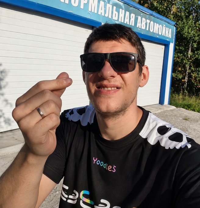 A man wearing sunglasses and a black t-shirt with 'yoodles' text and skull graphics makes a finger-heart gesture in front of a building with Russian signage 'АВТОМОЙКА'.