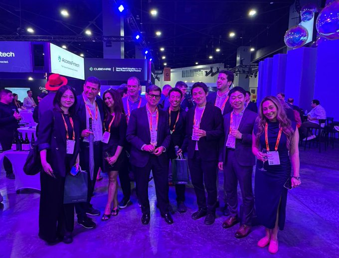 A group photo of professionals wearing conference badges posing together at what appears to be a tech or industry event, with purple and blue lighting illuminating the venue which displays sponsors like "cobeWORKS" and "Acossystems."