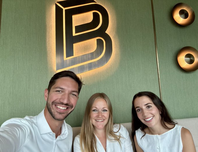 Three smiling people pose for a selfie in front of a green wall featuring a large illuminated 'B' logo and wall sconces.