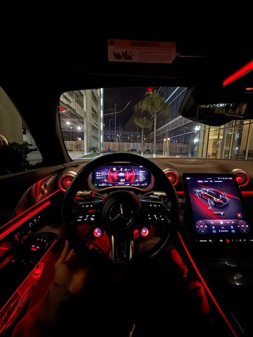 A nighttime view from the driver's seat of a Mercedes-Benz, showcasing its illuminated digital dashboard and steering wheel bathed in ambient red lighting. The luxurious interior contrasts with the post's casual "Good morning web3, Happy Sunday, $50 for breakfast" message, suggesting the driver is either heading out for or returning from an indulgent morning meal in style.
