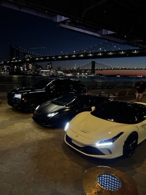 A nighttime photograph of luxury sports cars (including what appears to be a Rolls-Royce, Lamborghini, and Ferrari) parked with the illuminated Brooklyn Bridge and Manhattan skyline in the background.
