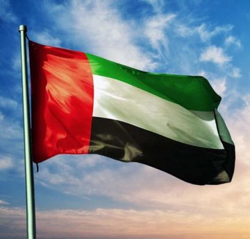 The national flag of the United Arab Emirates waving on a flagpole against a blue sky with clouds at sunset.