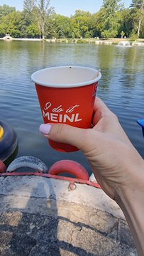 A hand holding a red paper cup with the text 'I do it MEINL' by a body of water.