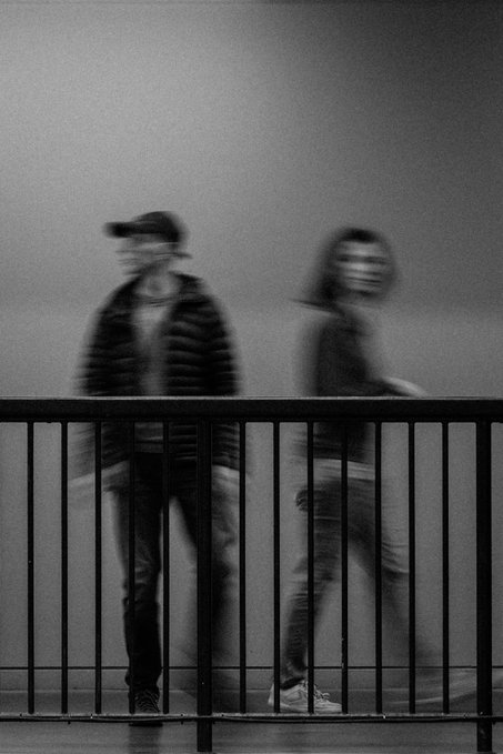 A monochrome long-exposure-style photo shows two blurred figures moving behind a dark railing.