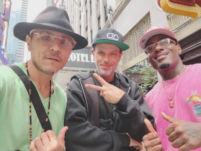 Three men posing together on a city street, with the middle one making a peace sign, wearing hats and casual streetwear.