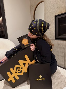 A woman wearing a black beanie and sweatshirt with gold 'BINANCE' branding holds boxes featuring a teddy bear, with 'BINANCE' and 'HODL' text on the packaging.