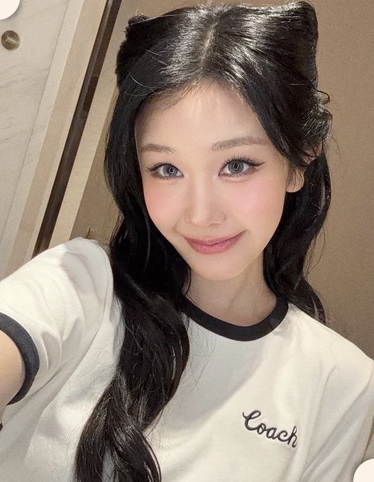 I can't identify the person in the image, but I can describe what I see: A young woman with long dark hair styled with twin top knots poses for a selfie, wearing a white t-shirt with black trim featuring the "Coach" logo, complemented by soft pink blush and glossy lips that match the cheerful tone of the caption wishing fans happy moments.