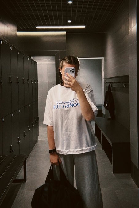 A person taking a mirror selfie in a locker room, wearing a white 'ECRU CLUB' t-shirt and grey pants while holding a phone and a black bag.