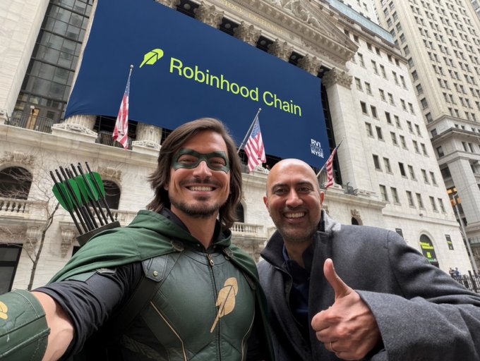 Two men pose for a selfie in front of the New York Stock Exchange, where a large screen displays "Robinhood Chain" alongside the company's feather logo. The man on the left is dressed in a Green Arrow costume with a mask and quiver of arrows, while the man on the right gives a thumbs-up—both celebrating Robinhood's blockchain initiative, as referenced in the caption "Two words: Robinhood Chain."