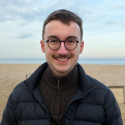 A smiling young man with short brown hair, glasses, and a mustache wears a dark puffy jacket over a brown zip-up sweater on a sandy beach.