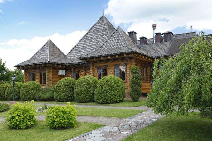 A traditional wooden log house with a dark shingled roof, surrounded by manicured spherical bushes and a stone pathway in a green lawn.
