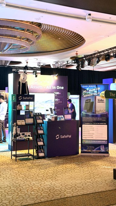 A SafePal cryptocurrency booth ("Assets - All In One") at an indoor conference or exhibition, featuring promotional materials and the text "DOUHAUS HARDWARE WALLET".