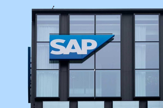 The blue and white 'SAP' logo displayed on a sign attached to a modern glass office building against a light blue sky.
