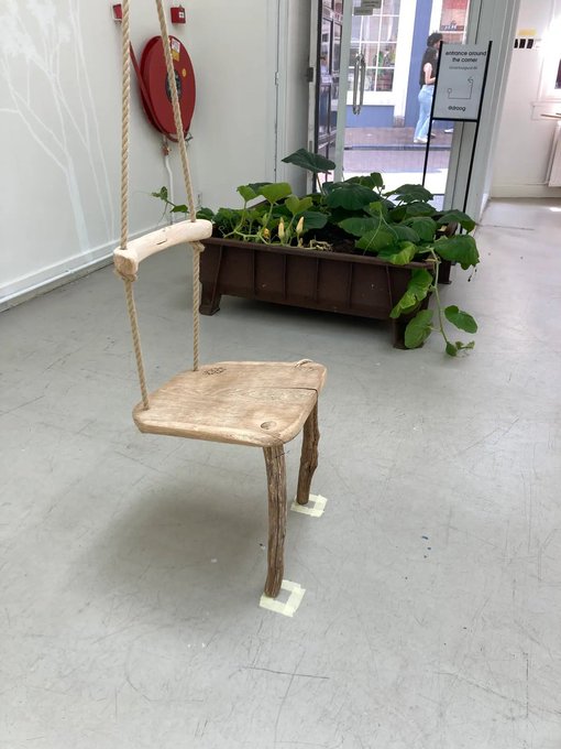 A rustic wooden swing suspended by rope hangs in an indoor space with a gray floor, near a planter box filled with green plants.