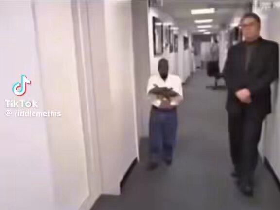 The image shows a man holding a book, possibly a cookbook or a recipe book, as he walks down an office hallway with another person following him. The hallway appears to be part of an office building, suggesting that they might be in a professional setting. The man carrying the book seems to be trying to sell it to someone else who is walking behind him.