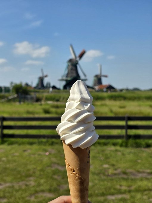 A hand holds up a tall swirl of soft-serve ice cream in a waffle cone, with traditional Dutch windmills and a green field blurred in the background under a blue sky.