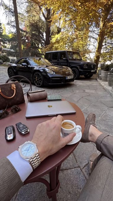 A first-person view shows a person enjoying an espresso at an outdoor café, with their hand resting on a saucer, wearing a luxury steel watch, a light blue dress shirt, and brown loafers. The table holds a closed MacBook with the Apple logo, a Louis Vuitton wallet, sunglasses, a brown monogram handbag, and two car key fobs, while a black Porsche 911 Turbo (license plate "34 DB 506") and a black Mercedes-Benz G-Class are parked on a stone driveway framed by autumn trees in the background. The image perfectly complements the motivational caption by visually showcasing the tangible rewards—luxury vehicles, designer accessories, and a refined lifestyle—that come from dedicated effort and hard work, serving as aspirational proof that ambition paired with action leads to success.