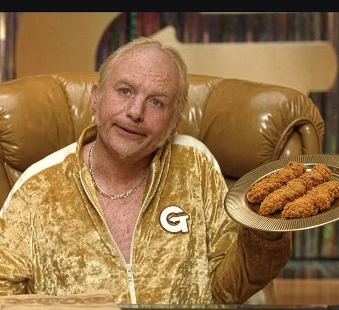 A man wearing a gold velour tracksuit with a 'G' logo sits on a leather couch holding a plate of fried chicken sticks, likely from a TV commercial.