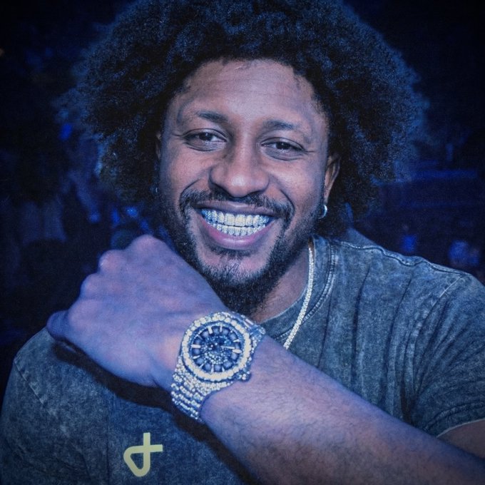 A smiling man with an afro and diamond-encrusted grill flashes a fully iced-out luxury watch against a blue-lit background, flexing his wrist to showcase the bling in line with his "gm (get money) big week" caption celebrating financial success.