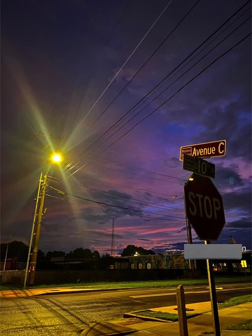 The image features a street scene at night with a stop sign next to an empty road. The sky above has a beautiful purple hue, creating a serene atmosphere. There are no people or cars visible in the photo, giving it a peaceful ambiance. The stop sign is located on the right side of the image and stands out against the dark background.