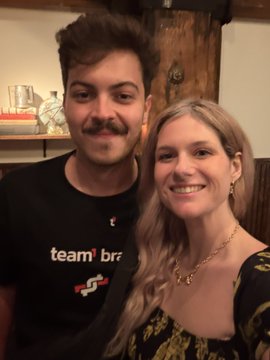 A man with a mustache wearing a black 'team brz' t-shirt standing next to a smiling woman with long blonde hair in a floral dress.