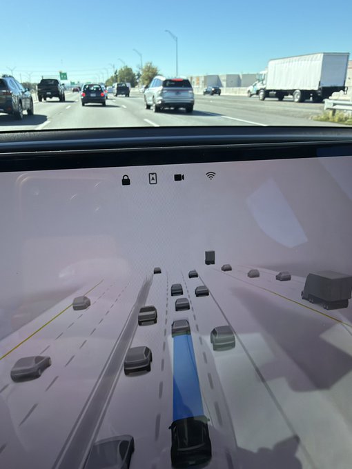 A dashboard view from inside a Tesla vehicle showing a 3D visualization of surrounding traffic on a highway under a clear blue sky.