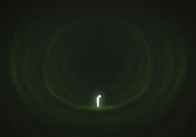 A small figure stands illuminated in the center of a dark space, surrounded by faint green circular light patterns.