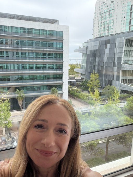 A selfie of a smiling woman taken through glass, with modern glass office buildings and greenery visible in the background.
