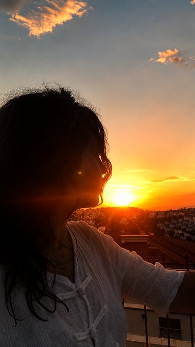 A woman with long dark hair is silhouetted against a vibrant golden sunset overlooking a cityscape, her face partially lit as she gazes into the distance. The image complements the post by visually conveying a reflective, travel-inspired moment—capturing the "journey continues" sentiment amid the author's busy summer and travels with a host family.