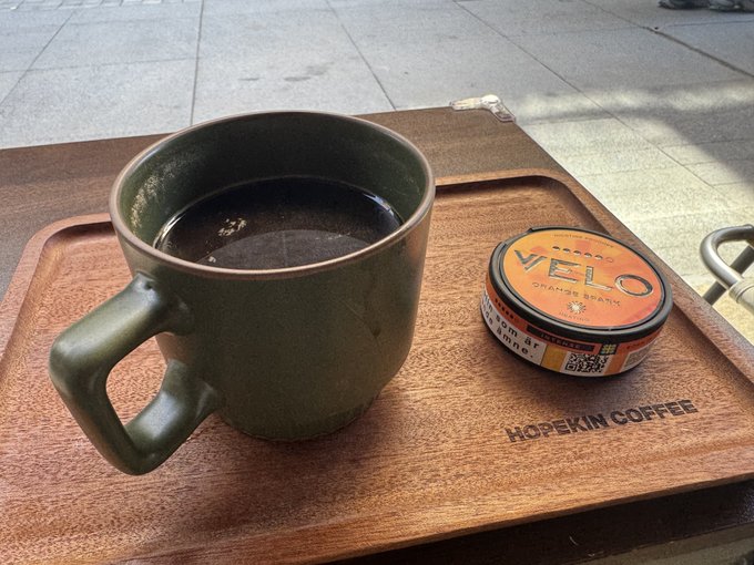 A green ceramic mug filled with black coffee sits on a wooden tray labeled "HOPEKIN COFFEE," accompanied by a round container of "VELO Orange Spark" nicotine pouches.
