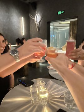 A group of friends clinks cocktail glasses together in a toast at a dimly lit bar or lounge with metallic countertops and ambient lighting.