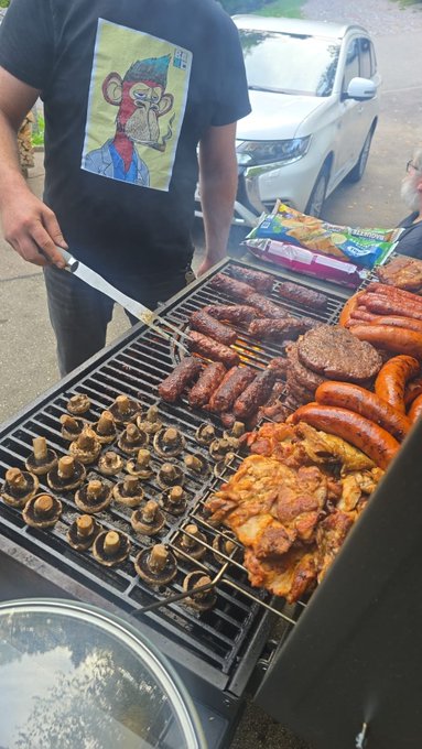 A person in a black Bored Ape t-shirt grilling a large assortment of meats and mushrooms on a barbecue outdoors.