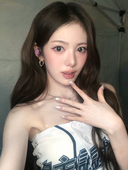 A woman with long dark hair wearing a white and navy cheerleader-style top, posing with her hand near her face.