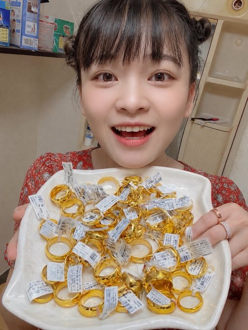 The image features a young woman holding up a white plate filled with gold rings, smiling for the camera. The plate contains several rings of varying sizes and designs, creating an eye-catching display. The background appears to be a kitchen or dining area, suggesting that this could be a special occasion or celebration involving these gold rings.