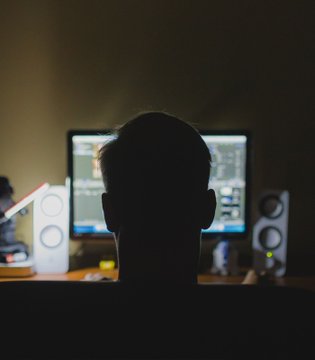 The image shows a person's head, with their back turned towards the camera, sitting behind a desk that has a computer monitor and speakers on it. The person appears to be focused on something on the screen of the monitor or possibly playing music through the speakers. The background is dark, which emphasizes the silhouette of the person against the light source from the monitor or speakers. The image does not provide any information about the content displayed on the monitor or speakers, but it seems that the person might be engaged in some form of entertainment or work-related activity.