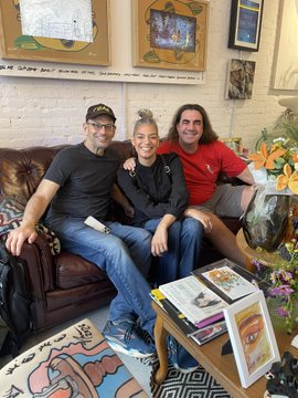 Three people sit together on a brown leather couch in a room decorated with colorful artwork, plants, and patterned rugs.