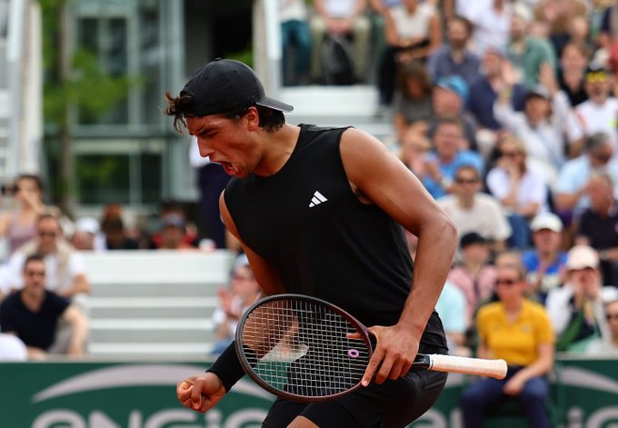 A tennis player wearing a black Adidas cap and sleeveless shirt pumps his fist in celebration while gripping his racket at the Cincinnati Open, with spectators visible in the stands behind him. The image visually represents the Marcos Giron match referenced in the post, with the player's passionate celebration capturing the intensity of the qualifying tournament contest against Henrique Rocha.